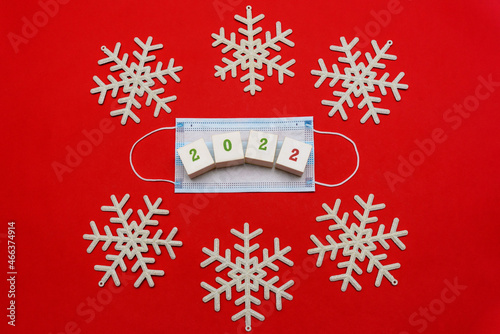 A medical mask with an inscription from cubes 2022 on a red background with snowflakes form a circle. Top view with copy space, space for text. Celebrating the New Year. Happy New Year 2022 concept.