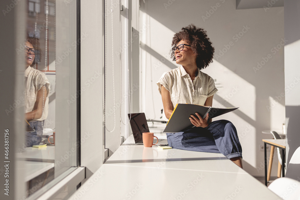 Happy woman in glasses sitting on the table while holding notebook and looking out the window Happy woman in glasses sitting on the table while holding notebook and looking out the window