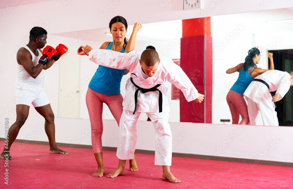 Asian woman exercising joint lock move with her trainer in kimono, African-american man boxing in front of mirror behind them.