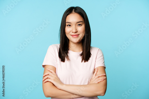 Smiling asian girl in t-shirt, holding arms crossed, ready for action pose, standing over blue background