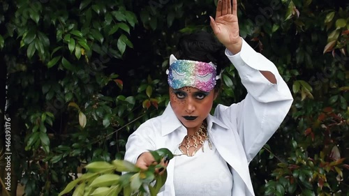 Latin woman in disguise with a make-up face and a colorful mask on her forehead making movements with her arms and facial expressions in a nature background