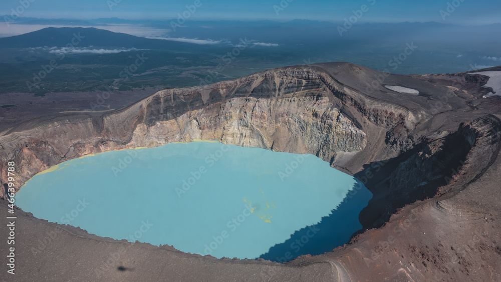 There is a turquoise lifeless acid lake in the crater of the volcano. Sulfurous deposits are visible on the water. Steep rocky shores. The shadow of a helicopter on the slope. Aerial view. Kamchatka