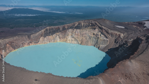 There is a turquoise lifeless acid lake in the crater of the volcano. Sulfurous deposits are visible on the water. Steep rocky shores. The shadow of a helicopter on the slope. Aerial view. Kamchatka