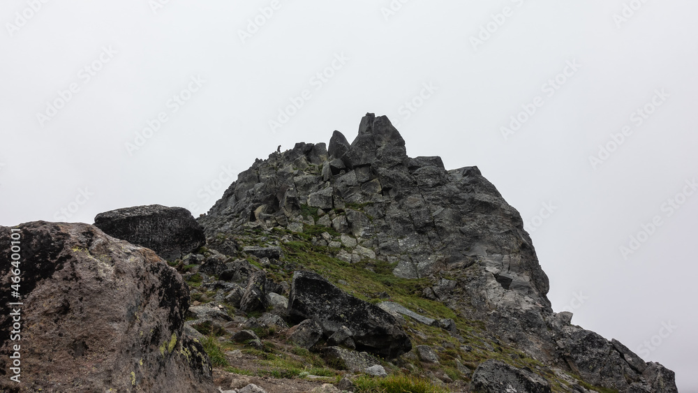The top of the cliff with bizarre outlines against the foggy sky. Cracks on the rocks and sparse vegetation. A tiny silhouette of a man on a peak is visible. Kamchatka. Mount Camel