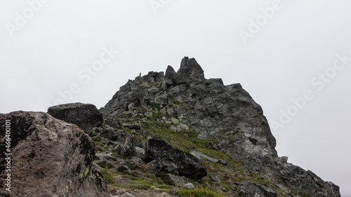 The top of the cliff with bizarre outlines against the foggy sky. Cracks on the rocks and sparse vegetation. A tiny silhouette of a man on a peak is visible. Kamchatka. Mount Camel © Вера 