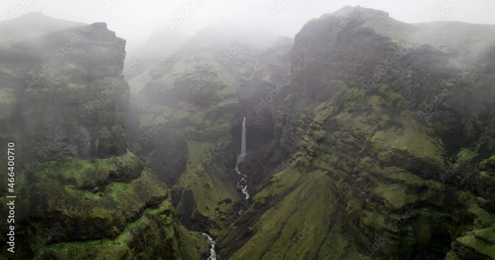 A mossy canyon with a waterfall in the background during a moody day. A river flows at its centre. Located in Iceland.