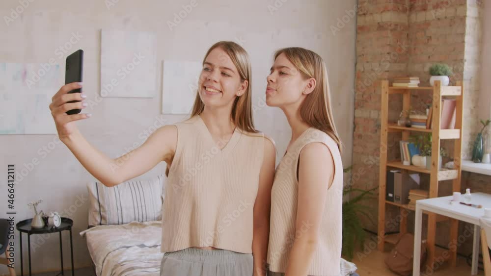 Handheld medium shot of happy young twin girls standing in their bedroom and taking selfie on mobile phone, then looking at photo Handheld medium shot of happy young twin girls standing in their bedroom and taking selfie on mobile phone, then looking at photo