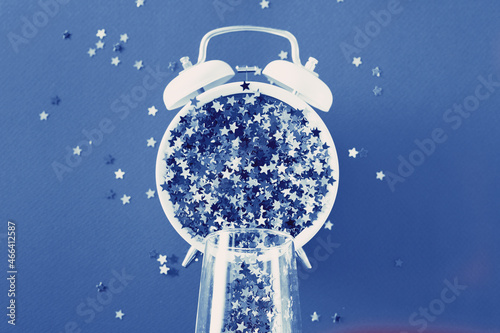 glass with sequins, alarm clock and star shaped confettie on blue background. photo