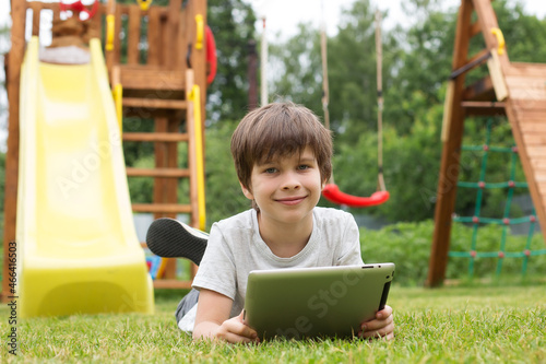 a a smiling boy with a tablet is sitting on the green grass on the playground. Online training, online games