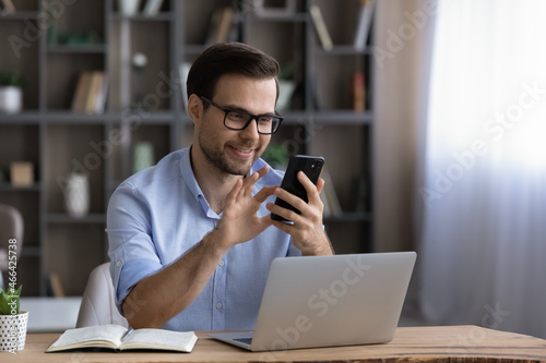 Happy handsome young man in eyewear looking at cellphone screen, reading message, or email, communicating distantly with client or playing games distracted from computer work in modern office.