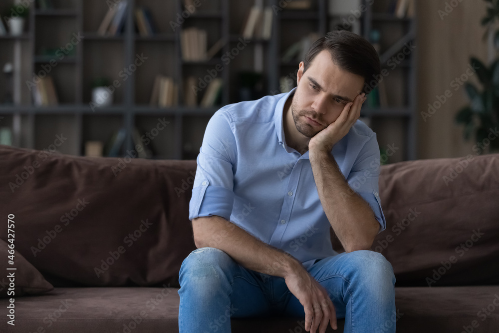 Bored millennial generation man sitting on sofa, feeling lonely at home. Unhappy young male suffering from negative thoughts, having depressive mood or problems in relations, regretting mistakes. Bored millennial generation man sitting on sofa, feeling lonely at home. Unhappy young male suffering from negative thoughts, having depressive mood or problems in relations, regretting mistakes.