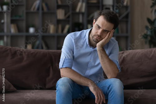 Bored millennial generation man sitting on sofa, feeling lonely at home. Unhappy young male suffering from negative thoughts, having depressive mood or problems in relations, regretting mistakes.