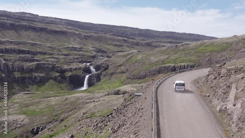 White car travels on dirt road passing scenic waterfall in Iceland, aerial photo