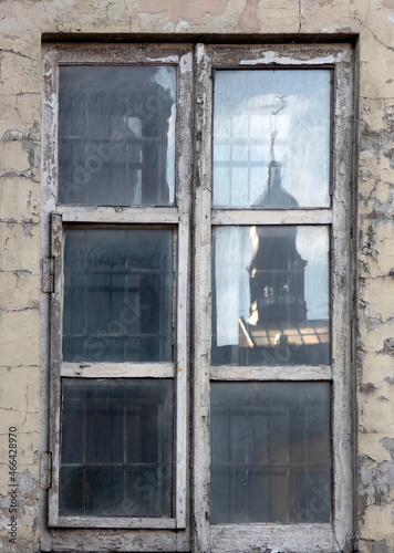 A window with bars in the wall of a very old house with peeling paint. One part of the window reflects the sunlit tower of an old church with a rooster on a spire.
