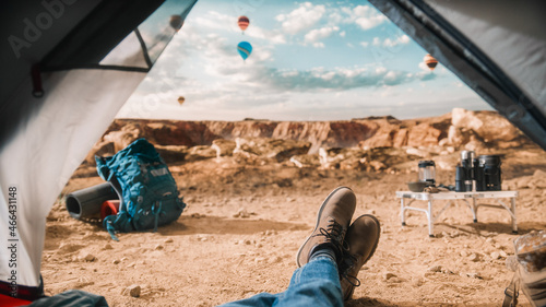 POV View of a Tourist Resting in a Tent Outdoors on Top of a Rocky Mountain with Canyon Valley and Flying Hot Air Balloons in the Sky. Backpacker Wearing Hiking Boots and Dusty Traveller Jeans. 