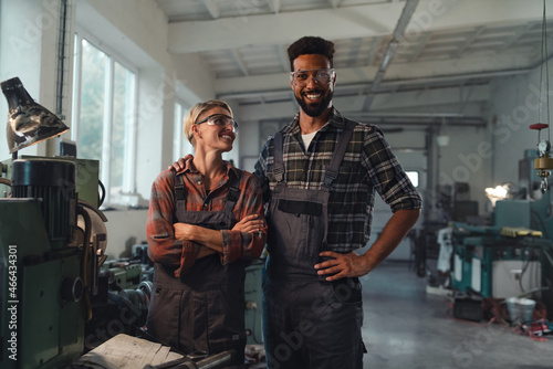 Portrait of young biracial industrial colleagues working indoors in metal workshop, smiling and looking at camera.