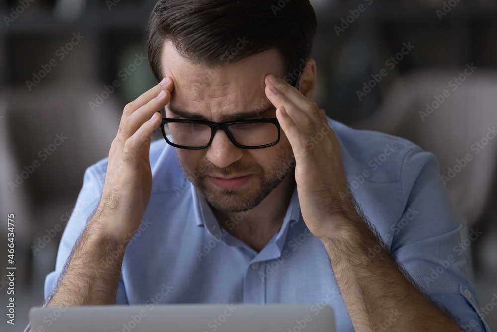 Head shot frustrated young male manager worker in eyeglasses massaging temples suffering from sudden severe headache or high blood pressure, overworking on computer, feeling unhealthy unwell in office Head shot frustrated young male manager worker in eyeglasses massaging temples suffering from sudden severe headache or high blood pressure, overworking on computer, feeling unhealthy unwell in office