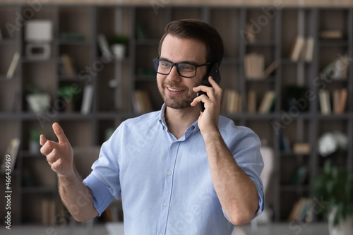 Happy young businessman in eyewear holding pleasant phone call conversation, discussing good project news with partners, giving professional consultation to client, communicating distantly in office.
