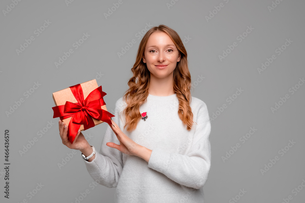 Young happy woman with blonde curly hair posing on gray background with gift box decorated with red ribbon. New Year Women's Day birthday holiday concept Young happy woman with blonde curly hair posing on gray background with gift box decorated with red ribbon. New Year Women's Day birthday holiday concept