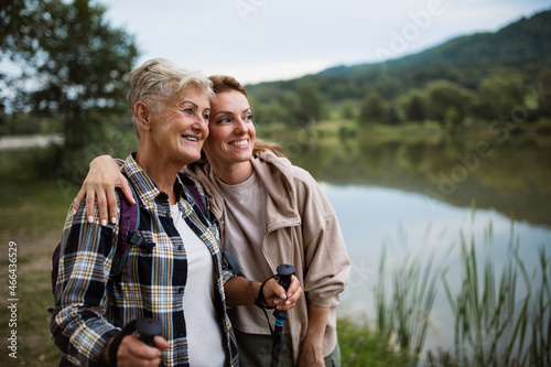 Happy senior mother hiker embracing with adult daughter when looking at lake outdoors in nature