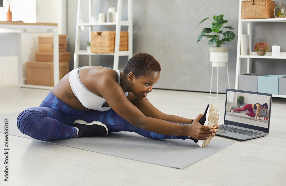 Happy young woman does regular sports workouts at home. Positive beautiful black lady doing leg stretching exercise while having an online class with her fitness instructor on her modern notebook PC - obrazy, fototapety, plakaty 