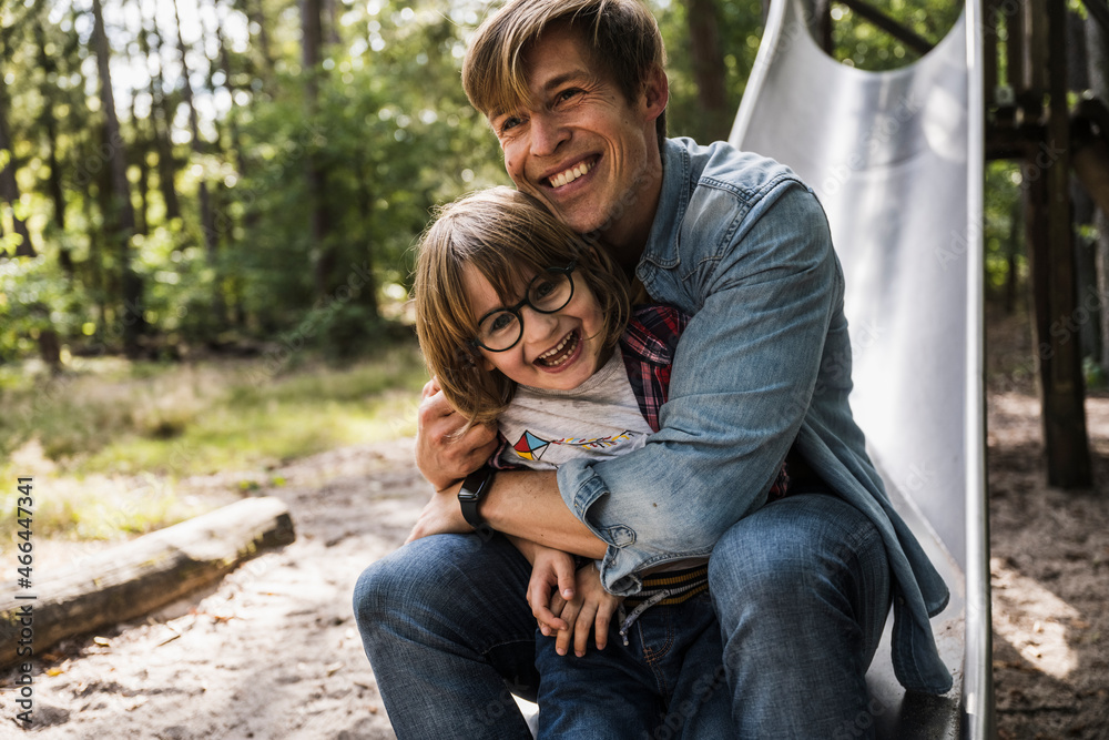 Father hugging son while sitting on slide in forest Father hugging son while sitting on slide in forest