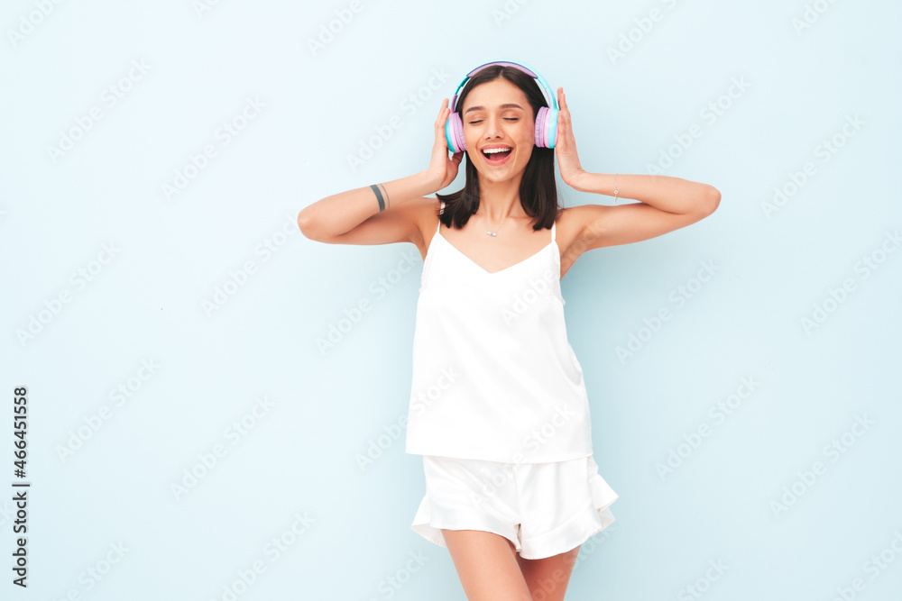 Beautiful smiling woman dressed in white pajamas. Sexy carefree model listening music in wireless headphones. Adorable and positive female posing near light blue wall in studio.Cheerful and happy Beautiful smiling woman dressed in white pajamas. Sexy carefree model listening music in wireless headphones. Adorable and positive female posing near light blue wall in studio.Cheerful and happy