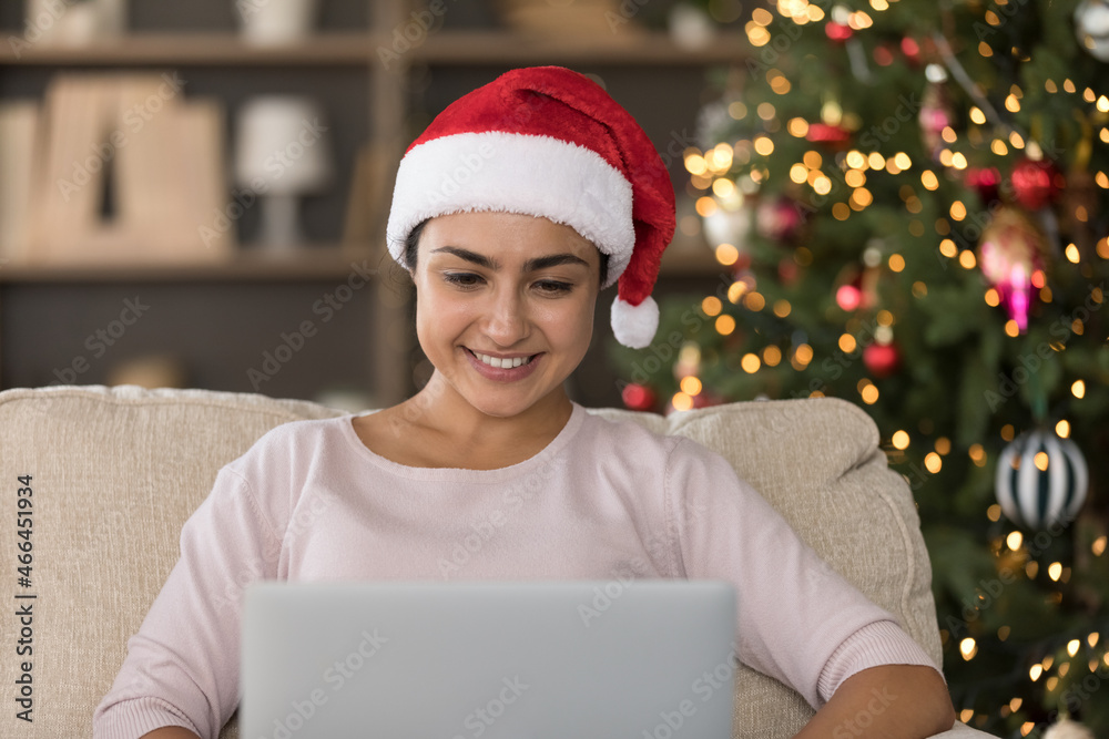 Smiling millennial Indian woman in red Santa hat looking at laptop screen, watching funny video, reading greetings with New Year in social network, celebrating Christmas miracle time online at home. Smiling millennial Indian woman in red Santa hat looking at laptop screen, watching funny video, reading greetings with New Year in social network, celebrating Christmas miracle time online at home.