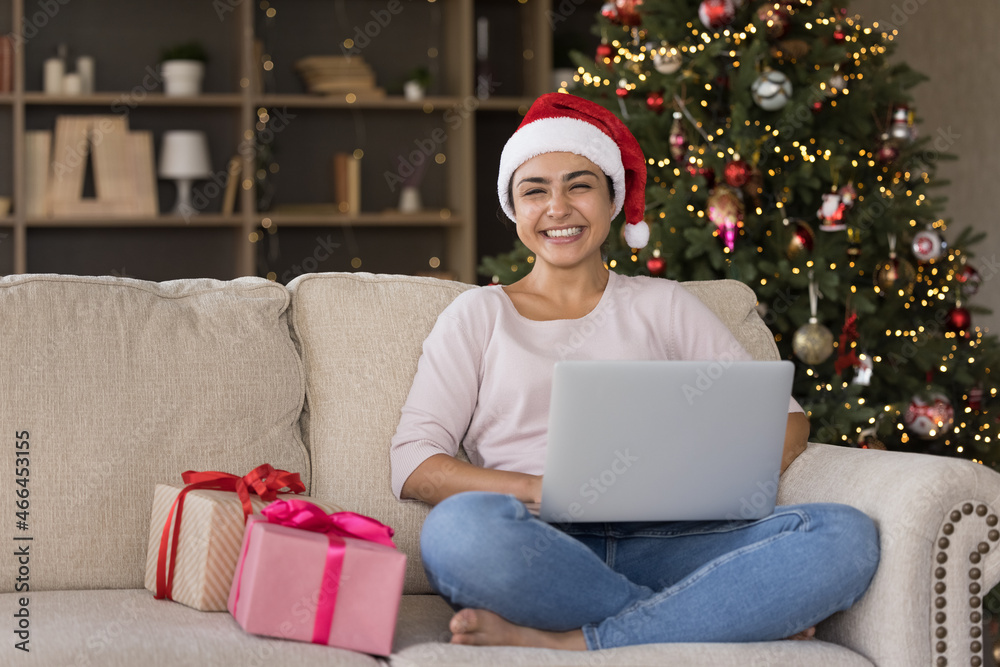 Happy millennial Indian woman in festive red hat holding computer on laps, looking at camera feeling excited sitting on couch with wrapped Christmas gifts near decorated tree, New Year celebration. Happy millennial Indian woman in festive red hat holding computer on laps, looking at camera feeling excited sitting on couch with wrapped Christmas gifts near decorated tree, New Year celebration.
