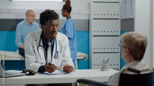 Black medic talking to disabled patient with handicap at desk in healthcare office. African american doctor helping elder woman with disability sitting in wheelchair. Old person at checkup photo