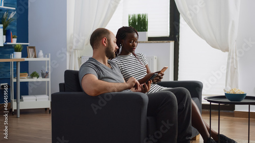 Interracial people using smartphones while sitting together at home. Multi ethnic couple holding devices and chatting on couch in living room. Modern mixed race lovers with technology