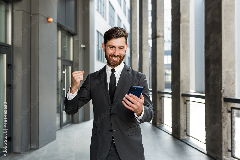 Successful and happy businessman shouts with joy and satisfaction of winning, reads good news from a mobile phone, outside office looks at camera Successful and happy businessman shouts with joy and satisfaction of winning, reads good news from a mobile phone, outside office looks at camera