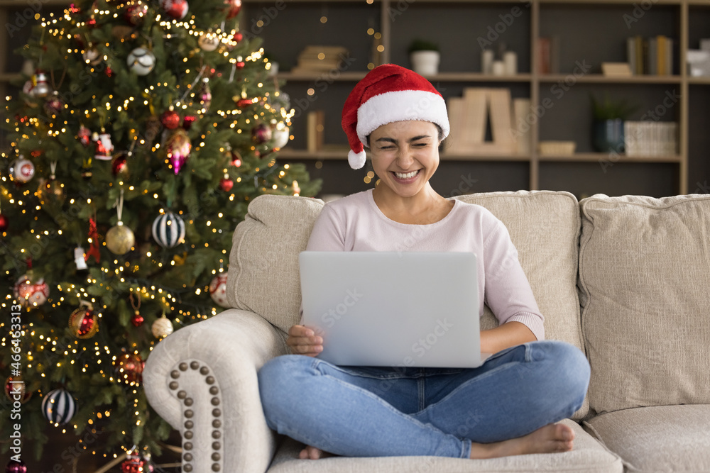 Happy millennial Indian woman in festive red hat holding computer on laps, laughing reading Christmas wishes or watching funny New Year movies online, resting on comfortable couch holidays celebration