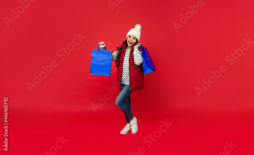 glad child in winter clothes hold shopping bags on red background with copy space, closeout