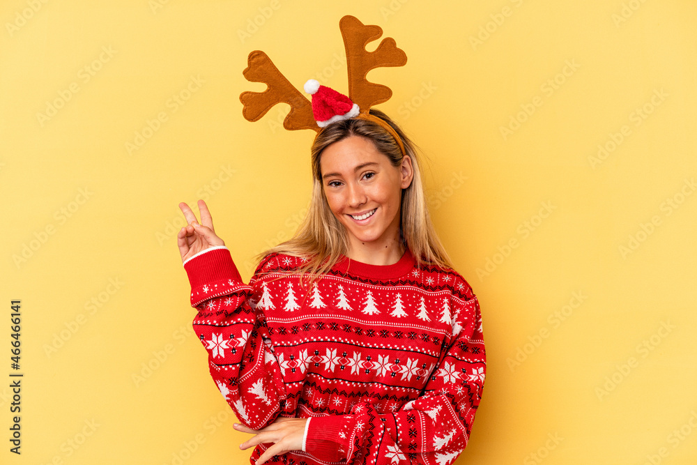 Young caucasian woman wearing a christmas reindeer hat isolated on beige background joyful and carefree showing a peace symbol with fingers. Young caucasian woman wearing a christmas reindeer hat isolated on beige background joyful and carefree showing a peace symbol with fingers.