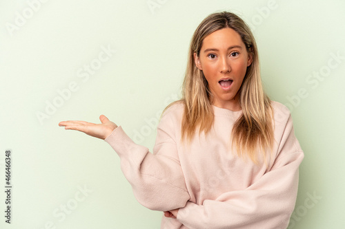 Young caucasian woman isolated on green background impressed holding copy space on palm.