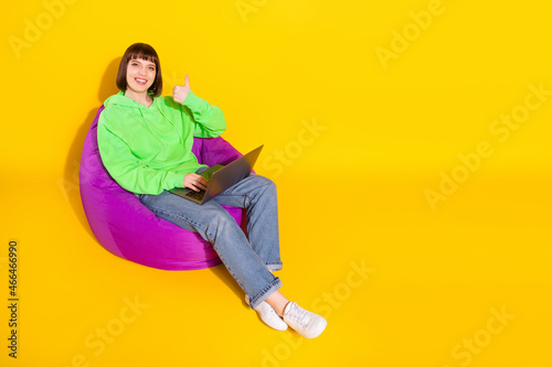 Portrait of pretty cheerful girl sitting in chair using laptop showing thumbup isolated over bright yellow color background © deagreez