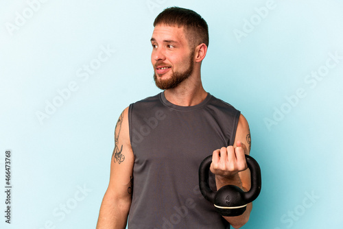 Young caucasian man holding ketlebell isolated on blue background looks aside smiling, cheerful and pleasant. photo