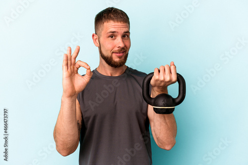 Young caucasian man holding ketlebell isolated on blue background cheerful and confident showing ok gesture. photo