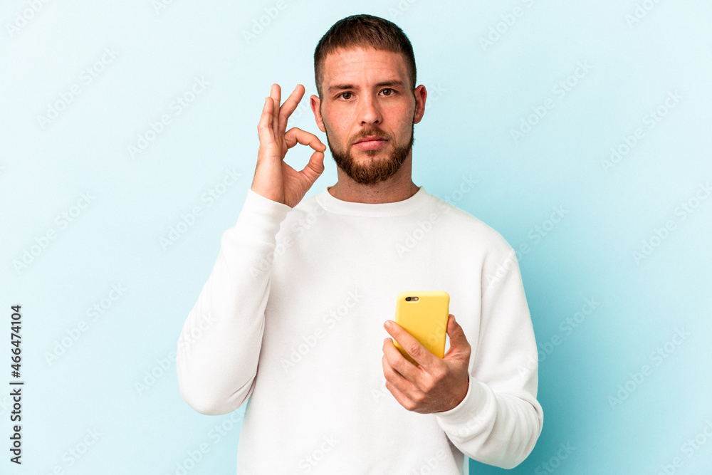 Young caucasian man holding mobile phone isolated on blue background with fingers on lips keeping a secret. Young caucasian man holding mobile phone isolated on blue background with fingers on lips keeping a secret.