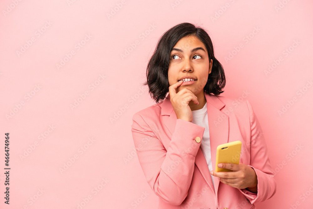 Young latin woman holding mobile phone isolated on pink background relaxed thinking about something looking at a copy space. Young latin woman holding mobile phone isolated on pink background relaxed thinking about something looking at a copy space.