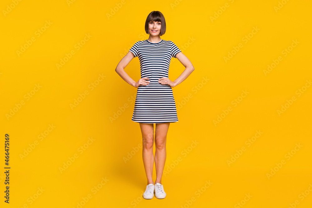 Full length photo of cute brunette millennial lady stand wear striped dress sneakers isolated on yellow background