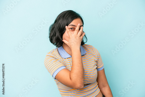 Young latin woman isolated on blue background blink at the camera through fingers  embarrassed covering face.