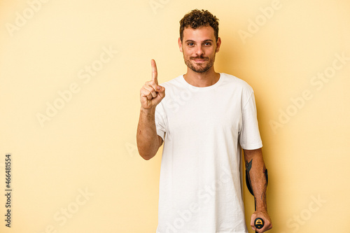 Young caucasian man holding crutch isolated on yellow background showing number one with finger. photo