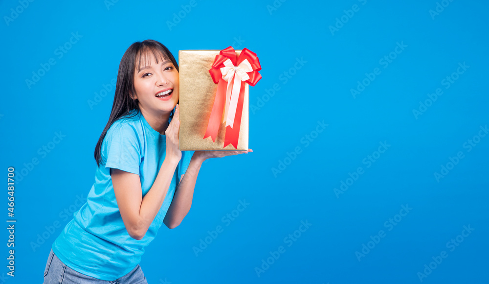 Surprise greeting anniversary birthday or celebration christmas and new year. Cheerful excited young woman holding gold gift box with red ribbon in own hand while standing over isolated background Surprise greeting anniversary birthday or celebration christmas and new year. Cheerful excited young woman holding gold gift box with red ribbon in own hand while standing over isolated background