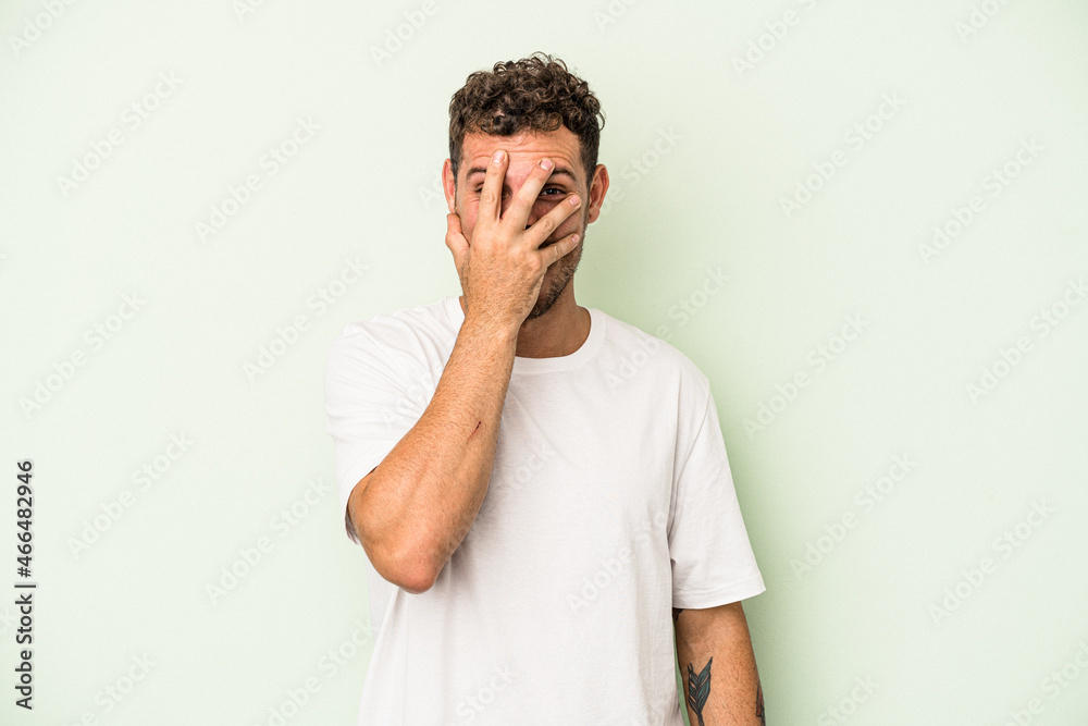 Young caucasian man isolated on green background blink at the camera through fingers, embarrassed covering face. Young caucasian man isolated on green background blink at the camera through fingers, embarrassed covering face.