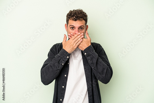 Young caucasian man isolated on green background covering mouth with hands looking worried.