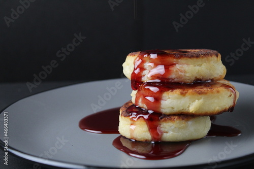 food breakfast cheesecakes curd pancakes three pieces lie stack of slides on top of each other and pour jam sauce cherry red on a gray plate on a black background