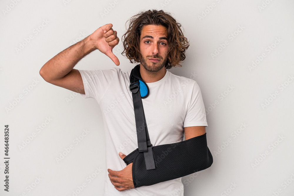 Young caucasian man with broken hand isolated on gray background showing a dislike gesture, thumbs down. Disagreement concept. Young caucasian man with broken hand isolated on gray background showing a dislike gesture, thumbs down. Disagreement concept.
