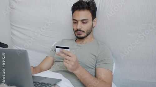 Middle Eastern guy is shopping online using laptop and credit card exressing happiness in bed. Finance and youth lifestyle concept. photo